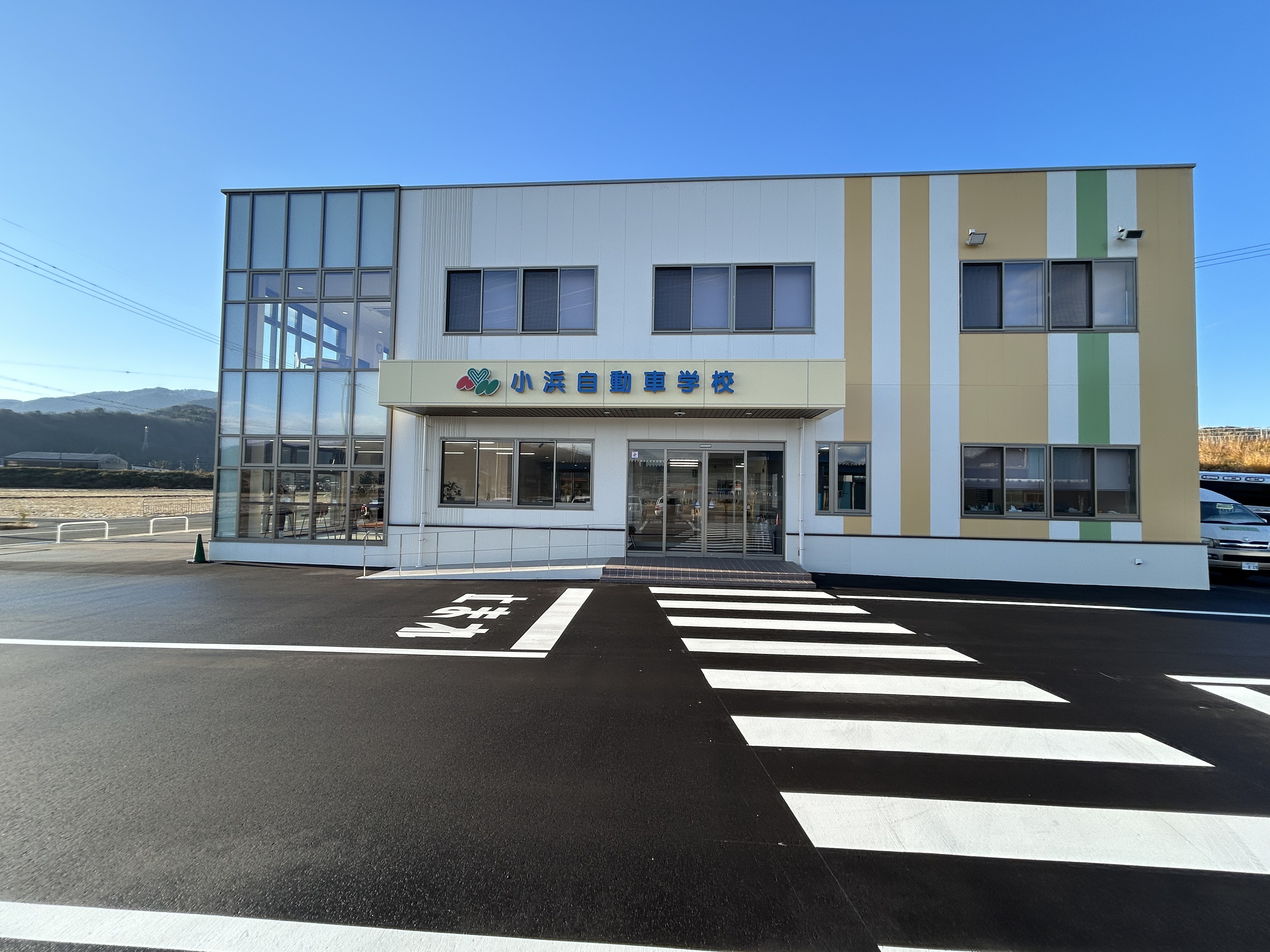 青空の下に建つ、白とクリーム色の外壁に緑の帯が入った2階建ての校舎。手前に横断歩道の白線がある